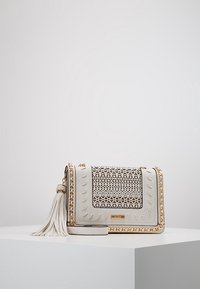 Bílá kožená crossbody taška s vyřezávaným vzorem, zlatými řetízkými akcenty, detaily s třásněmi a texturovanými stehy podél okrajů.