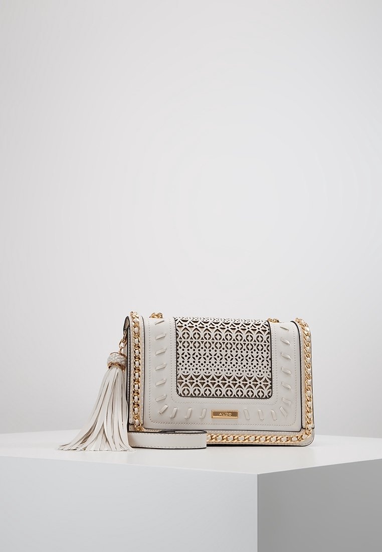 Bílá kožená crossbody taška s vyřezávaným vzorem, zlatými řetízkými akcenty, detaily s třásněmi a texturovanými stehy podél okrajů.
