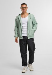 Světlá zelená bunda s kapucí a elastickými manžetami, nošená přes bílý tílko, v kombinaci s černými kalhotami a krémovými teniskami. Viditelné detaily strukturované tkaniny.