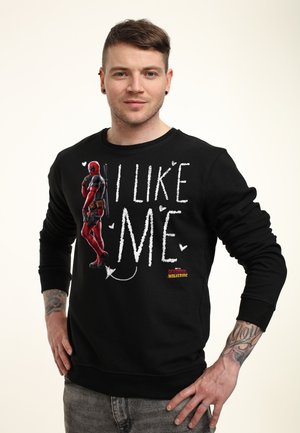 Homme tatoué portant un sweat-shirt noir avec un graphisme Deadpool et le texte « I Like Me », posant les mains sur les hanches devant un fond uni.