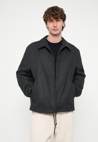 Sötétszürke gyapjúkabát gallérral, elején gombos lezárással és két oldalzsebbel. Az alsó szegélyén húzózsinór található az állítható illeszkedésért.