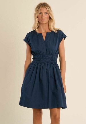Donna bionda che indossa un vestito blu navy a maniche corte con vita arricciata e scollo a V, in piedi contro uno sfondo beige.