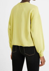 Keltainen puuvillainen collegeneule löysällä istuvuudella, resoripäinen pääntie ja pitkät hihat. Helmassa on hieman lyhennetty leikkaus takana.