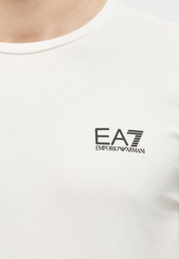 Κοντινό πλάνο λευκού μπλουζιού με στρογγυλή λαιμόκοψη και μαύρο λογότυπο "EA7 Emporio Armani" στην αριστερή περιοχή του στήθους.
