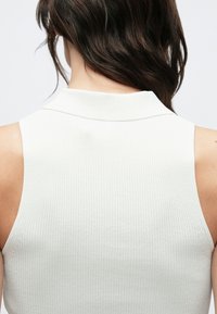 Priliehavý bezrukávový rebrovaný top v krémovej farbe s vysokým golierom. Hladká textúra a jemné švy pozdĺž výstrihu a ramenných švov.