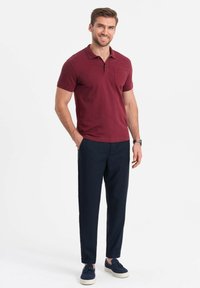 Polo shirt bordeaux con taschino, abbinato a pantaloni blu e scarpe blu con suola bianca. L'outfit ha un aspetto casual.