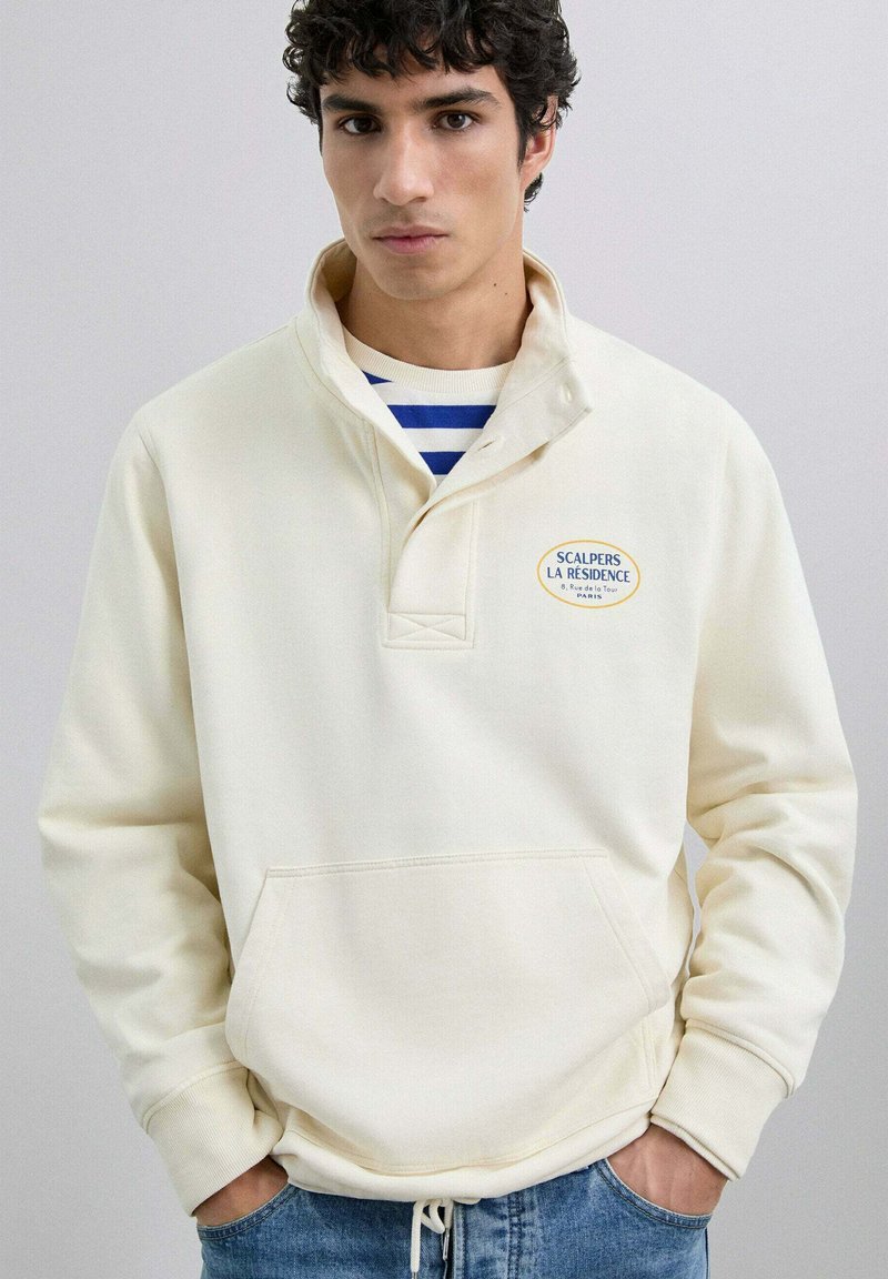 Sudadera de pullover crema con cuello de media cremallera, bolsillo frontal tipo pouch y logotipo bordado. Combinada con jeans azules. Textura suave.