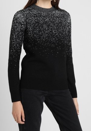 Woman wearing a black and gray speckled knit sweater with long sleeves and black pants against a plain background.