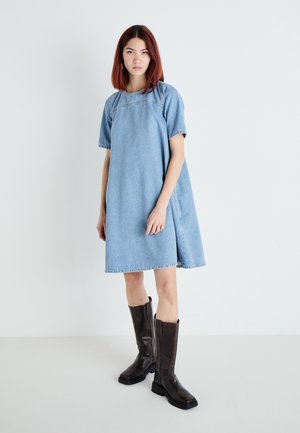 Light blue denim dress with short sleeves, relaxed fit, and contrast stitching. Paired with glossy brown knee-high boots.
