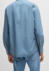 Camisa de manga larga de color azul claro con una textura suave, puños abotonados y un cuello clásico. La vista trasera revela un dobladillo redondeado.