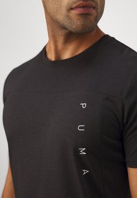 T-shirt preta feita de tecido macio, com decote redondo e uma impressão vertical do logo branco no lado esquerdo. Textura suave.