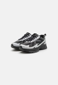 Sneakers featuring a mesh upper in silver and black, with a sturdy black rubber sole and textured accents. Black laces and pull tab noted.