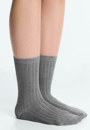 Ein Paar Beine trägt graue, gerippte Socken bis zur Mitte der Wade und steht auf einer weißen Fläche mit minimalem Hintergrund.