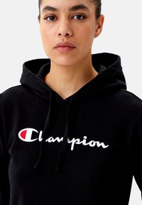 Černá mikina s kapucí vyrobená z bavlny s bílým vyšívaným logem "Champion" a červeným akcentem. S tkaničkami a texturovaným povrchem.