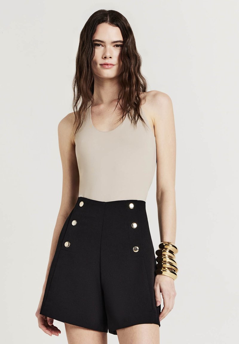 Beiger ärmelloser Bodysuit kombiniert mit hochtaillierten schwarzen Shorts, die mit goldenen Knopfakzenten versehen sind. Glatter Stoff und strukturierte Silhouette.