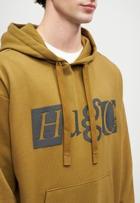 Olivgrüner Kapuzenpullover mit einer großen Kängurutasche vorne, Zugkordeln und einem großen, strukturierten schwarzen Logodruck auf der Brust. Weicher Stoff.