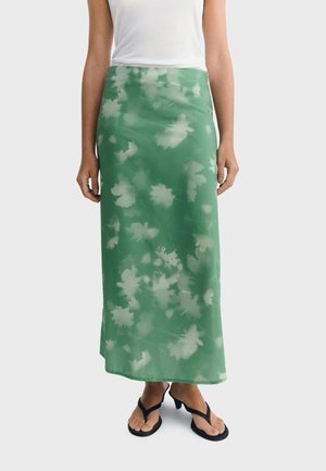 Femme portant une jupe midi tie-dye verte, un haut sans manches blanc et des sandales à talons noires, debout devant un fond uni.