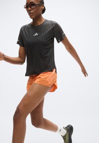 Chemise à manches courtes rayée noire avec le logo Adidas, associée à un short orange et des chaussures de sport noires. Pose de course, design et ajustement concentrés.