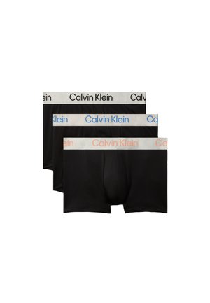 Hombre musculoso de pie descalzo con las manos detrás de la espalda, vistiendo calzoncillos bóxer negros de Calvin Klein sobre un fondo gris liso.