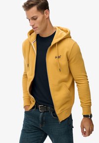 Uomo che indossa una felpa gialla senape con zip sopra una maglietta blu, jeans blu, cintura nera e orologio, che guarda in basso con le mani vicino alle tasche.