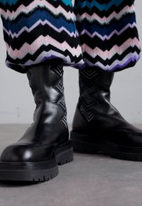Cizme de glezne negre din piele cu o talpă groasă și accente cu model zigzag pe părțile laterale. Purtați cu un material tricotat cu un model multicolor.