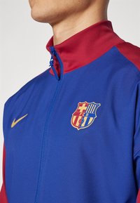 Röd och blå träningsjacka med hög krage, full dragkedja, Nike-swoosh samt FC Barcelonas klubbemblem i guld och livfulla färger.