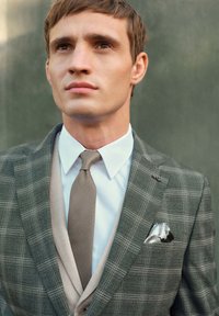 Completo grigio a quadri con rever a punta, camicia bianca, cravatta marrone testurizzata e fazzoletto da taschino argentato con dettagli a motivo. Texture del tessuto liscia.