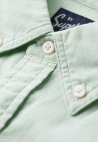 Lichtgroen overhemd met kraag, voorzien van twee witte knopen en textuurstof. Label zichtbaar met de tekst "Superdry". Close-up van het kraagdetail.
