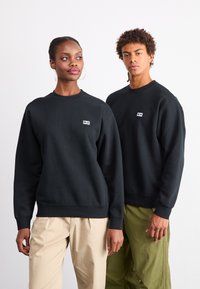 Mustat ümarad kaelusega dressid, millel on ribakudumiga varrukad ja alumine serv, ning rinnal väike logo. Mudel, kes kannab khaki pükse, seisab teise mudeli kõrval.