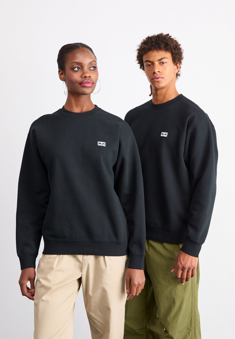 Mustat ümarad kaelusega dressid, millel on ribakudumiga varrukad ja alumine serv, ning rinnal väike logo. Mudel, kes kannab khaki pükse, seisab teise mudeli kõrval.