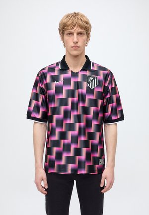 Un jeune homme se tient devant un fond uni et porte un maillot de football à carreaux noirs et roses de l’Atlético Madrid.