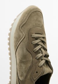 Light brown suede sneaker with a textured surface, lace-up design, perforated detail on the toe, and a chunky white rubber sole.