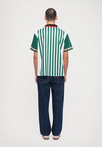 Korte mouwen shirt met groene en witte verticale strepen, zwarte kraag en mouwen met rode accenten. Gecombineerd met losse blauwe denim jeans.
