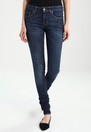 Kvinde iført mørkeblå slim fit jeans og sorte spidse flade sko, står med det ene ben krydset foran det andet.