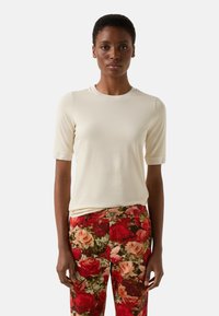 Krótki rękaw kremowo-beżowej bluzki z okrągłym dekoltem zestawiony z czerwonymi spodniami w kwiatowy wzór z różami w różnych kolorach na teksturowanym tle.