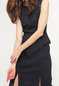 Musta pitsimekko, jossa on istuva bodice, aaltoileva helma ja sivureiät. Monimutkaiset brodeeratut kuviot luovat tekstuurista visuaalista yksityiskohtaa.