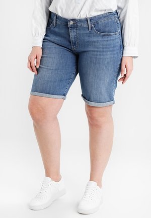 Personne portant un short en jean bleu avec ourlet roulé, une chemise blanche à manches longues et des baskets blanches, debout devant un fond blanc.
