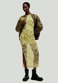 Oliven grønn bomberjakke over en gjennomsiktig, gul leopardmønstret kjole med sideslitt, parret med robuste svarte combat-boots.