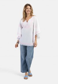 Tunic alb cu detalii brodate roz și dantelă, asortat cu blugi evazați de un albastru deschis și sandale albastre.
