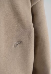 Beige long-sleeve top made of ribbed fabric featuring a subtle tonal logo embroidered on the left side. Smooth texture with a fitted shape.