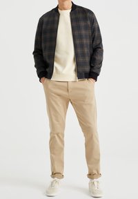 Muž v námořnickém a hnědém kostkovaném bomberu, krémovém svetru, béžových chinos kalhotách s ohrnutými nohavicemi a krémových teniskách stojí s rukama v kapsách.