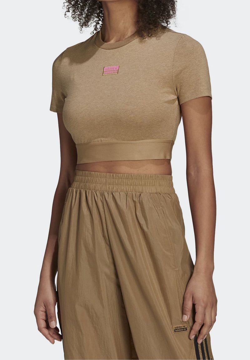 Crop top court beige à manches courtes avec un design ajusté et une texture lisse, doté d'une petite étiquette rose sur la poitrine, associé à un pantalon beige ample.
