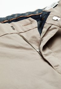 Nahaufnahme von beigen Hosen mit teilweise geöffnetem Metallreißverschluss, Knopfverschluss und sichtbarem blauen Denim-Bündchenfutter.