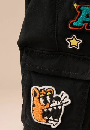 Tissu noir avec des patches texturés ; présente un personnage orange avec des accents verts et une figure en noir et blanc, ainsi qu'une étoile jaune.
