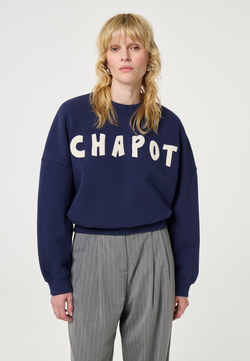 Felpa blu navy con la scritta "CHAPOT" in lettere crema. Presenta un collo a giro e spalle basse. Abbinata a pantaloni grigi a righe.