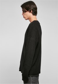 Suéter negro de punto con un corte holgado, que presenta una textura acanalada, mangas largas y un escote poco profundo. Usado sobre una camiseta negra.