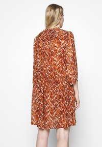 Femme aux cheveux blonds portant une robe longue jusqu'aux genoux à manches longues avec un imprimé animal abstrait rouge, orange et blanc, vue de dos.
