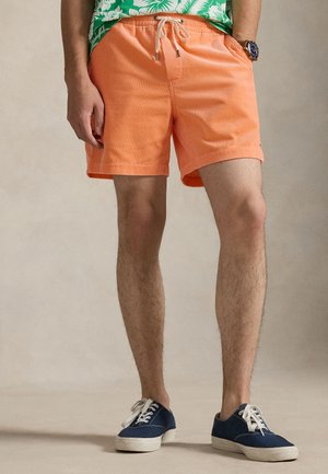 Narancssárga kordbársony rövidnadrág húzózsinóros derékkal és oldalzsebekkel, kék vászon tornacipővel párosítva, semleges háttér előtt.