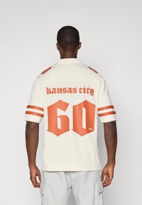 Kermajersey vaaleilla punaisilla yksityiskohdilla, jossa on painettuna "Kansas City" ja "60" tyylillä takana. Lyhyet hihat ja kauluksellinen kaula-aukko.