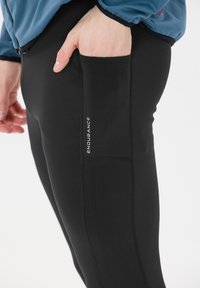 Leggings atléticos negros, hechos de tela elástica con corte ajustado, que cuentan con un bolsillo lateral y un pequeño logo de "ENDURANCE".